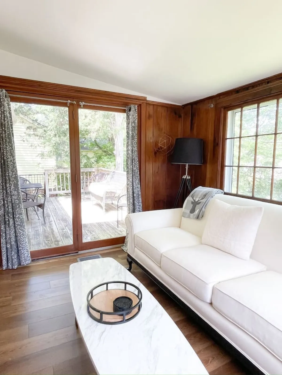 A cozy living room scene featuring a plush sofa and coffee table positioned near a large window, with warm sunlight filtering in and a soft lamp casting a gentle glow for evening relaxation. The arrangement suggests a comfortable spot for unwinding, sharing a drink, or connecting with loved ones.