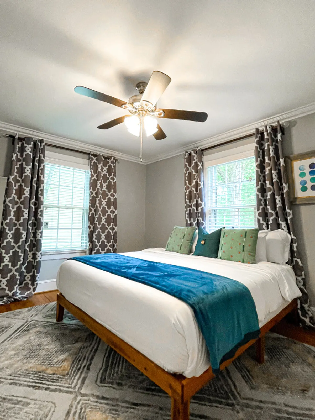 A serene bedroom with a king-size bed adorned with a turquoise throw, geometric-patterned curtains for privacy, a ceiling fan overhead, and a cozy reading area. The calming colors and soft linens evoke a sense of restfulness and comfort.