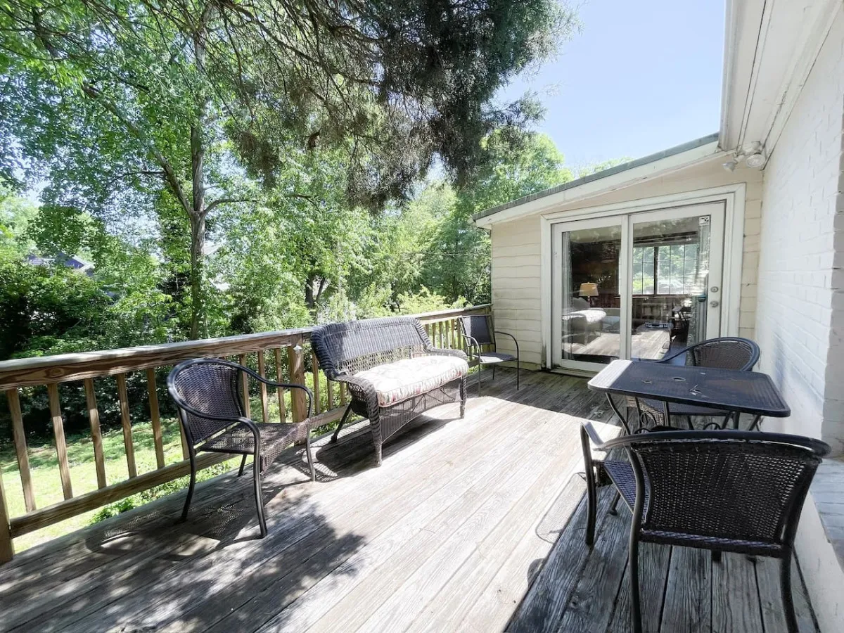 A welcoming exterior wooden deck adorned with wicker chairs, a cushioned bench, and tables, all surrounded by lush greenery. The setup suggests a serene place for outdoor dining, morning coffee, or social gatherings, with the natural backdrop hinting at neighborhood tranquility.