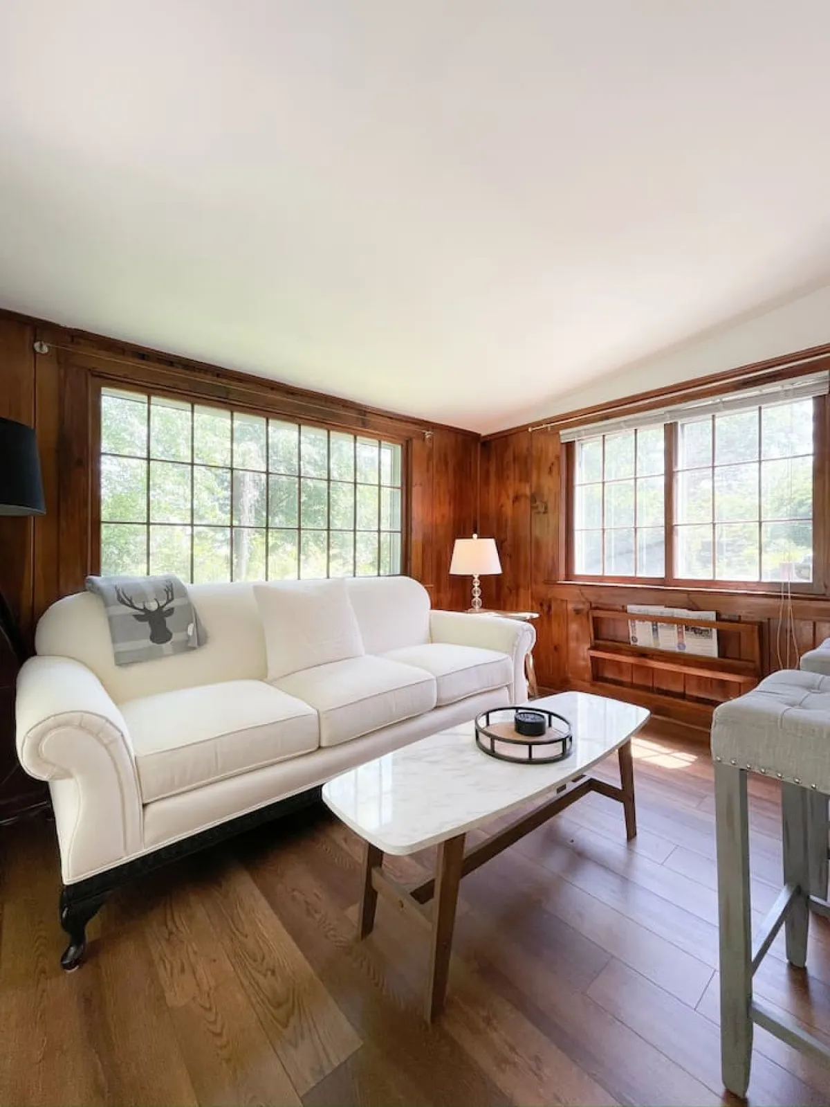 A spacious living room with a white sofa, coffee table, large windows that bathe the room in sunlight, and warm wood paneling. The space is styled for comfort, relaxation, and socializing, with ample room for guests or quiet personal time.