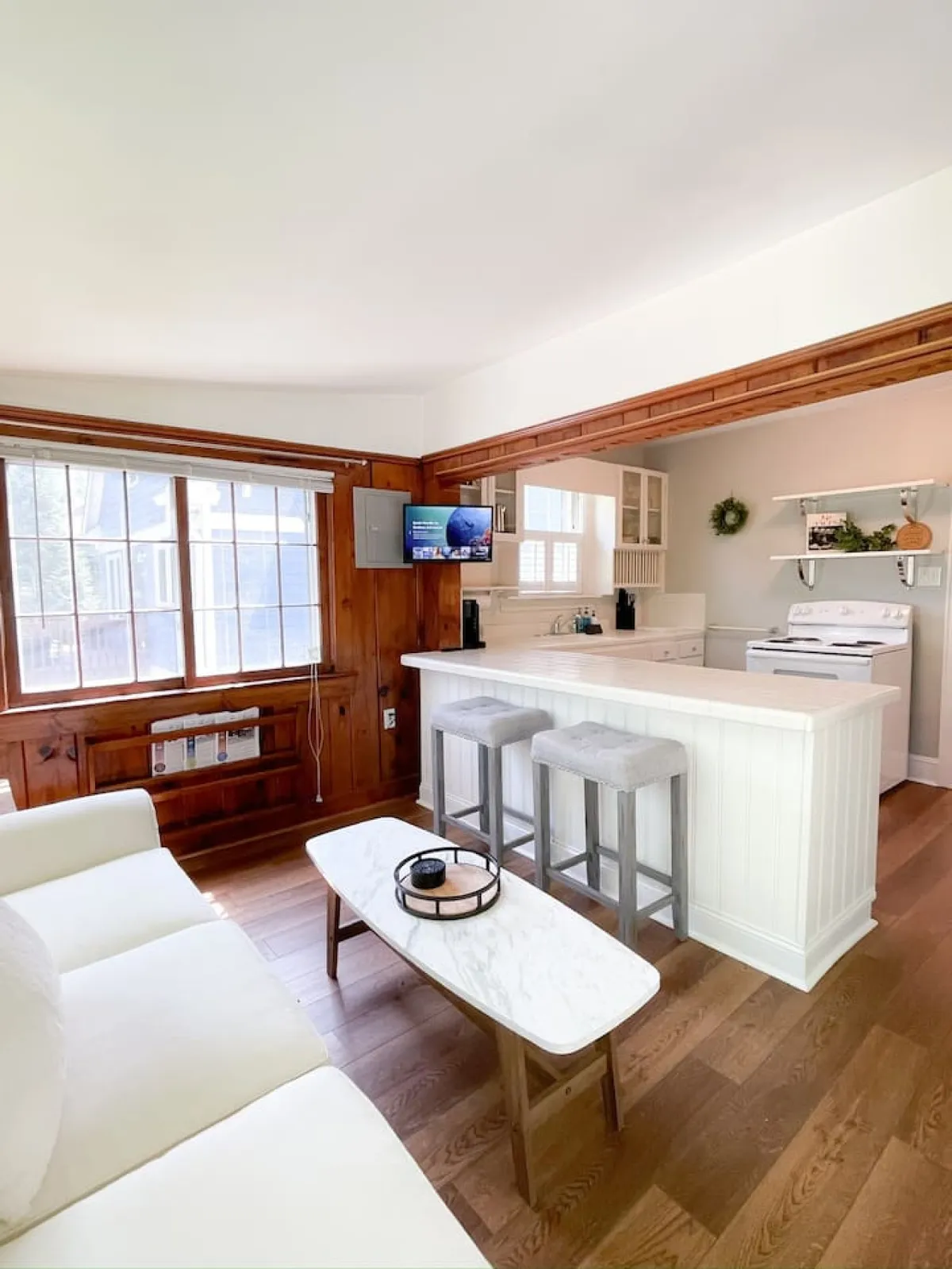 A cozy, open-plan living area featuring a comfortable sofa, a kitchen island with bar stools, a television, and a large window letting in natural light. The space feels both homely and modern, ideal for relaxing, entertaining, or just unwinding after a long day.