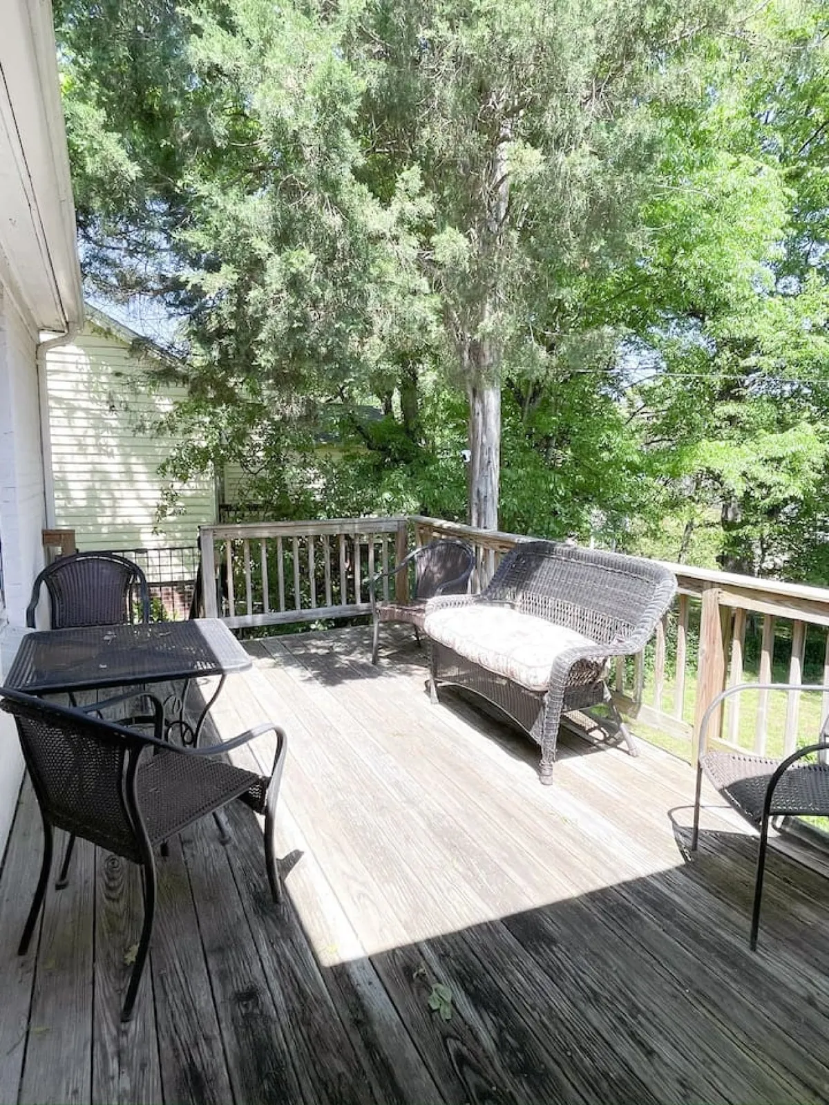 A tranquil exterior wooden deck set amidst abundant greenery, featuring comfortable wicker furniture. The scene exudes a calm, laid-back atmosphere ideal for unwinding after exploring the neighborhood, with hints of sunset or morning light filtering through the trees.