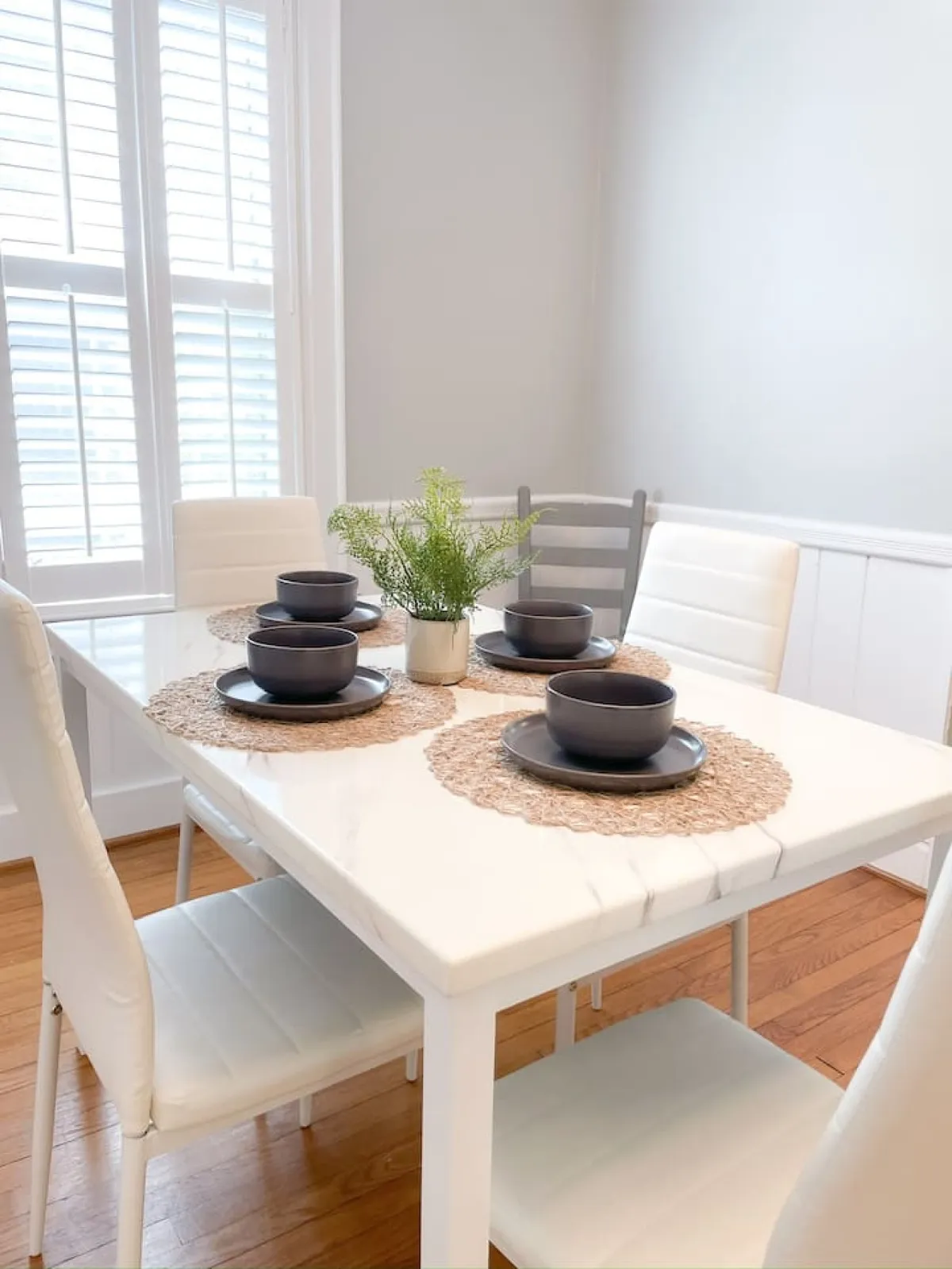 This image showcases a modern, intimate dining nook with a white marble table set for four, featuring black bowls, woven placemats, and a touch of greenery for warmth. The setting is perfect for shared meals, family dinners, or casual brunches, capturing the cozy, inviting atmosphere highlighted in the narrative.