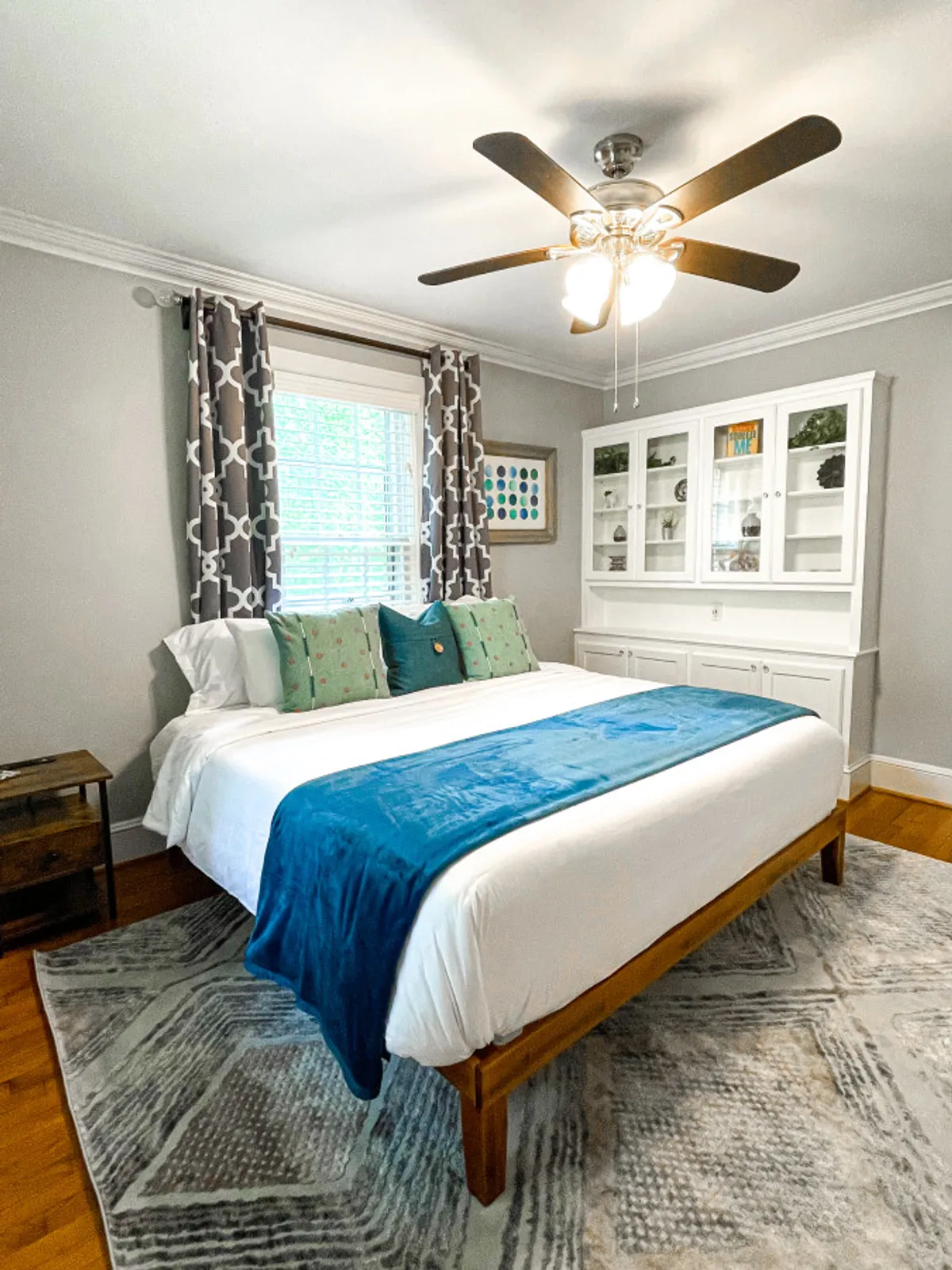 A tranquil bedroom featuring a comfortable bed, bedside table, window with curtains, and a display cabinet. The natural light filtering through the window and the subtle decor create a serene space ideal for unwinding.