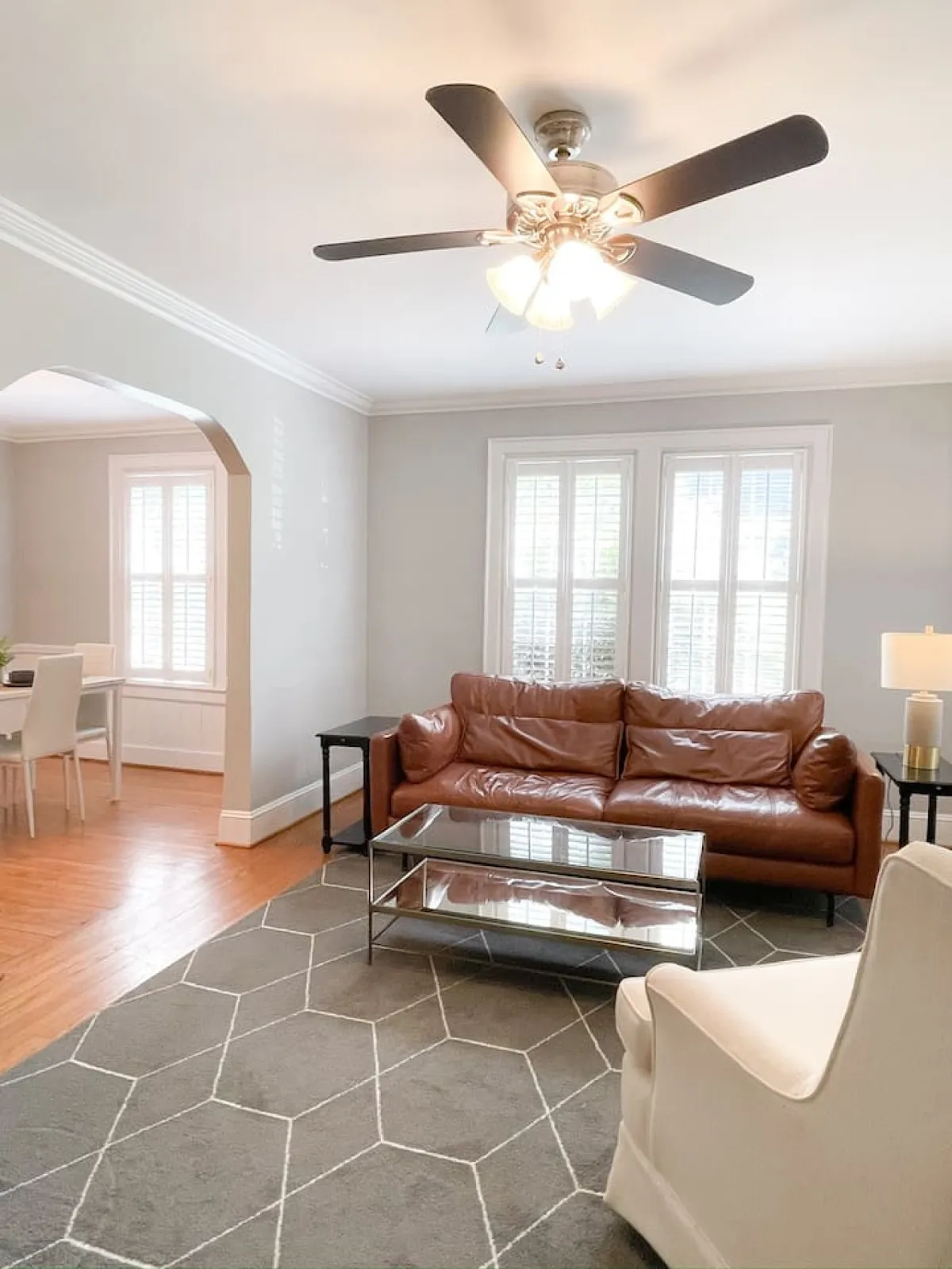 A bright, inviting space featuring a brown leather sofa, glass coffee table, white armchair, and ceiling fan. The arrangement is ideal for both relaxing solo or hosting friends and family, evoking a sense of ease and welcome.